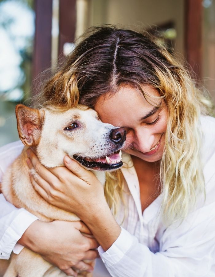 A smiling person with long hair embraces a tan dog